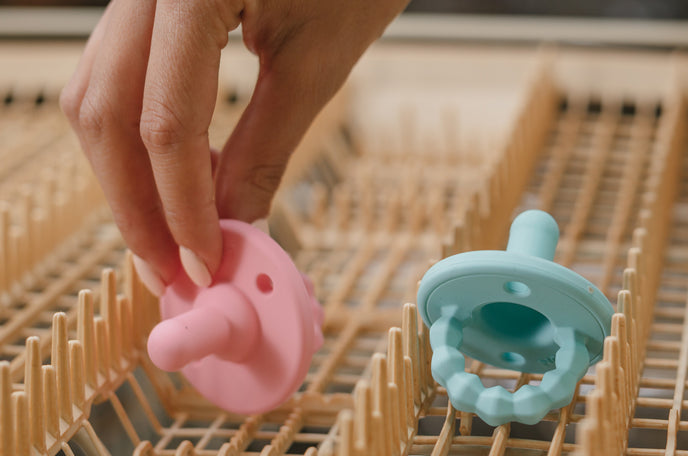 A pink and blue Nuby Silicone Softees Pacifier being placed on the top rack of a dishwasher.