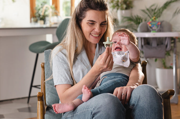 Smiling mother holding her child in her lap as he reaches for the Nuby pacifier in her right hand.