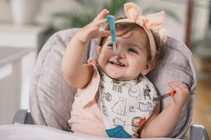 Happy baby girl eating a meal, playing with Nuby utensils in her highchair.