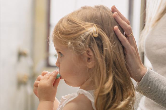 Toddler girl brushing her teeth with her mother's hand resting gently on the back of her head.