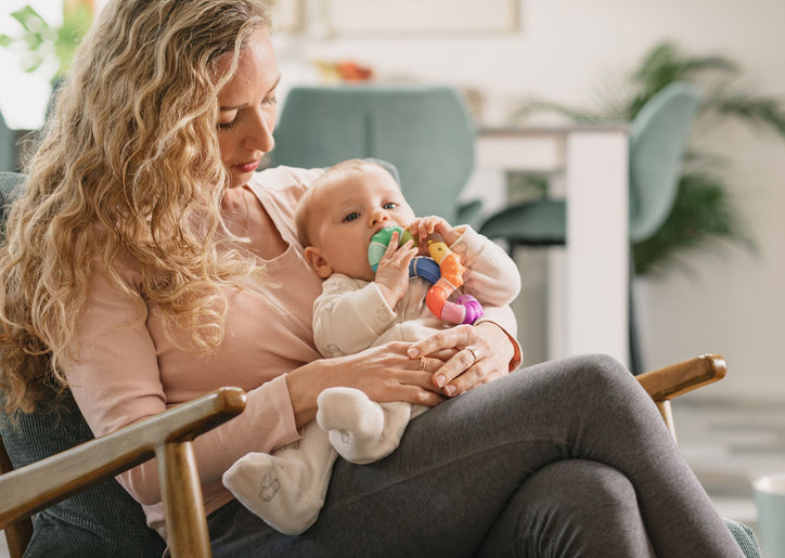 Woman holding a child in her lap. The child is teething, chewing on a Nuby Twista Rattle Teether Toy.