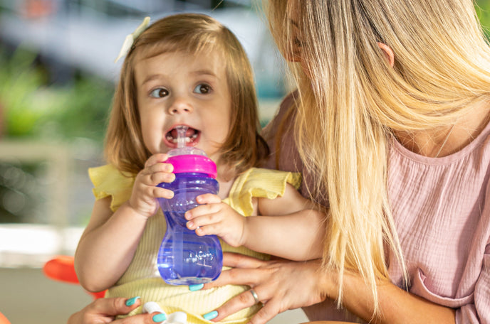 A young child next to her mother, learning to use a Nuby Flex Straw Leakproof Sippy Cup.