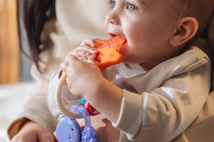 Infant using Nuby's IcyBite Sea Cooling Teether Keys while sitting on mother's lap.