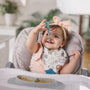 Happy baby girl eating a meal, playing with Nuby utensils in her highchair.
