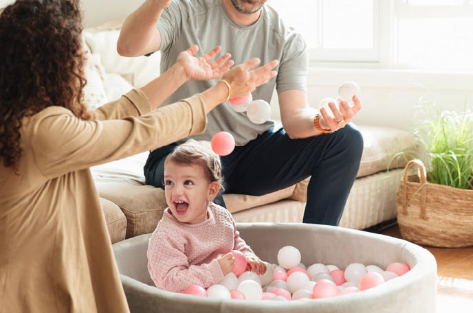 Happy toddler playing in gray Nuby ball pit with pink and white balls while smiling father and mother toss ball pit balls around.