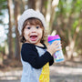 An excited child wearing a knitted cap outside, mouth agape, holding a purple and blue Nuby sippy cup with both hands.