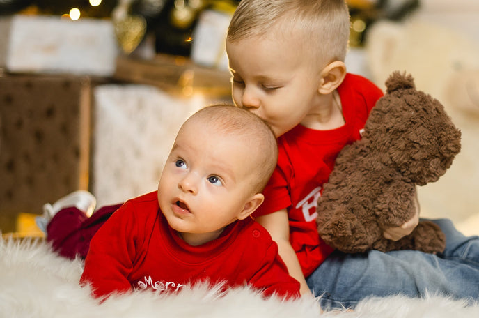 Two siblings in matching red shirts sitting on a plush, white rug. The older sibling is holding a teddy bear and kissing the top of the other's head.