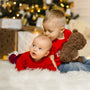 Two siblings in matching red shirts sitting on a plush, white rug. The older sibling is holding a teddy bear and kissing the top of the other's head.