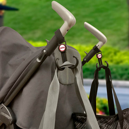 Close-up of two Nuby 360-Swivel Stroller Hooks (2 Pack) in pink attached to stroller handles, holding a grey strap and a black bag; green lawn and foliage blurred in the background.