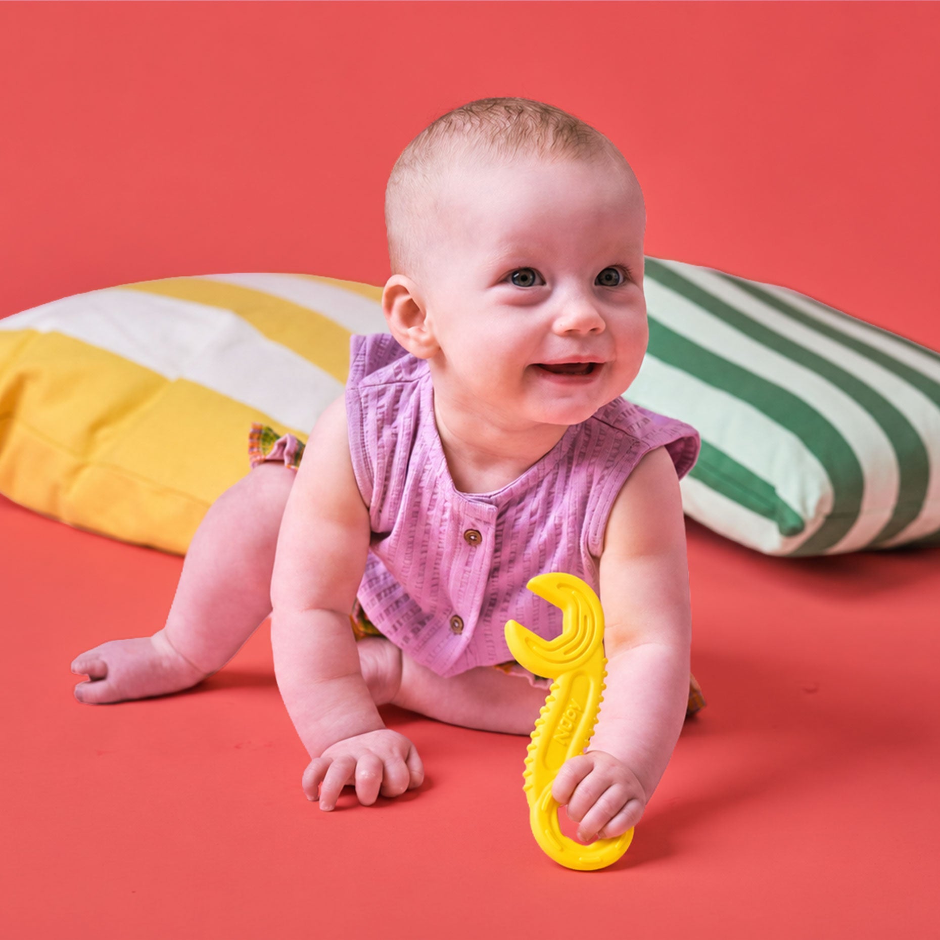 A smiling baby in a pink outfit sits on a red surface, holding a yellow Tool Teethers (4 Pack) teether shaped like a wrench. Two pillows—one with yellow and white stripes, the other green and white—are behind the baby.