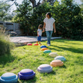 An adult and child walk hand in hand across StepPebbles and FREE Balance Board | Pastel, colorful foam stepping stones arranged on grass, perfect for indoor or outdoor play to help kids develop balance and coordination.
