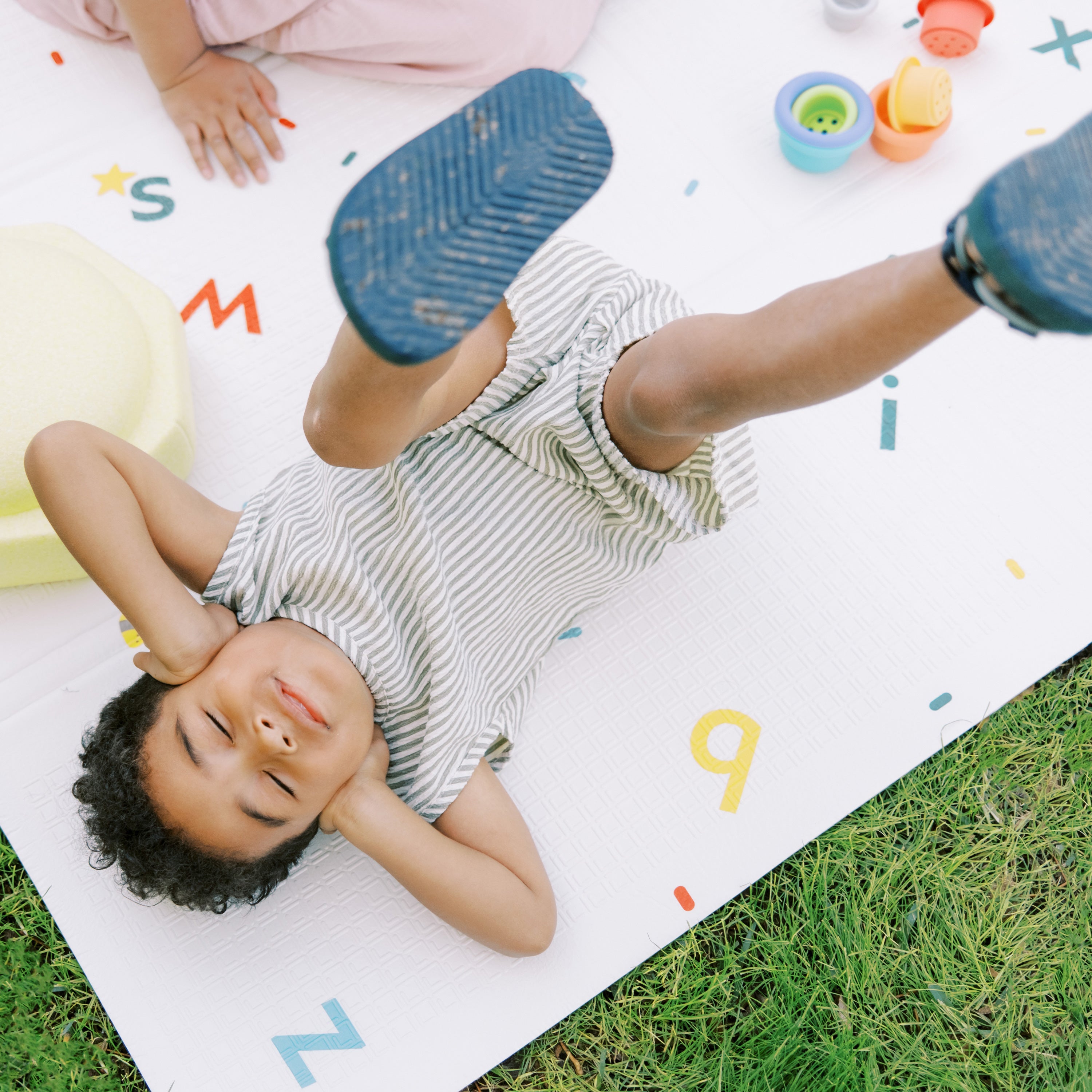 A child in a striped outfit smiles with closed eyes and raised legs while lying on the Reversible Floor Mat | Cars & Roads outdoors, surrounded by colorful toys and alphabet letters.