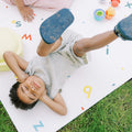 A child in a striped outfit smiles with closed eyes and raised legs while lying on the Reversible Floor Mat | Cars & Roads outdoors, surrounded by colorful toys and alphabet letters.