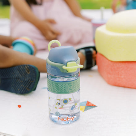 A Nuby US Thirsty Kids Flip-It BOLT Water Bottle | Space with a green and blue no-spill lid sits on a white outdoor surface, surrounded by children's hands and colorful toys.