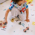 A young child in colorful clothes kneels on the Reversible Floor Mat | Cars & Roads, playing with wooden shapes and a toy car.