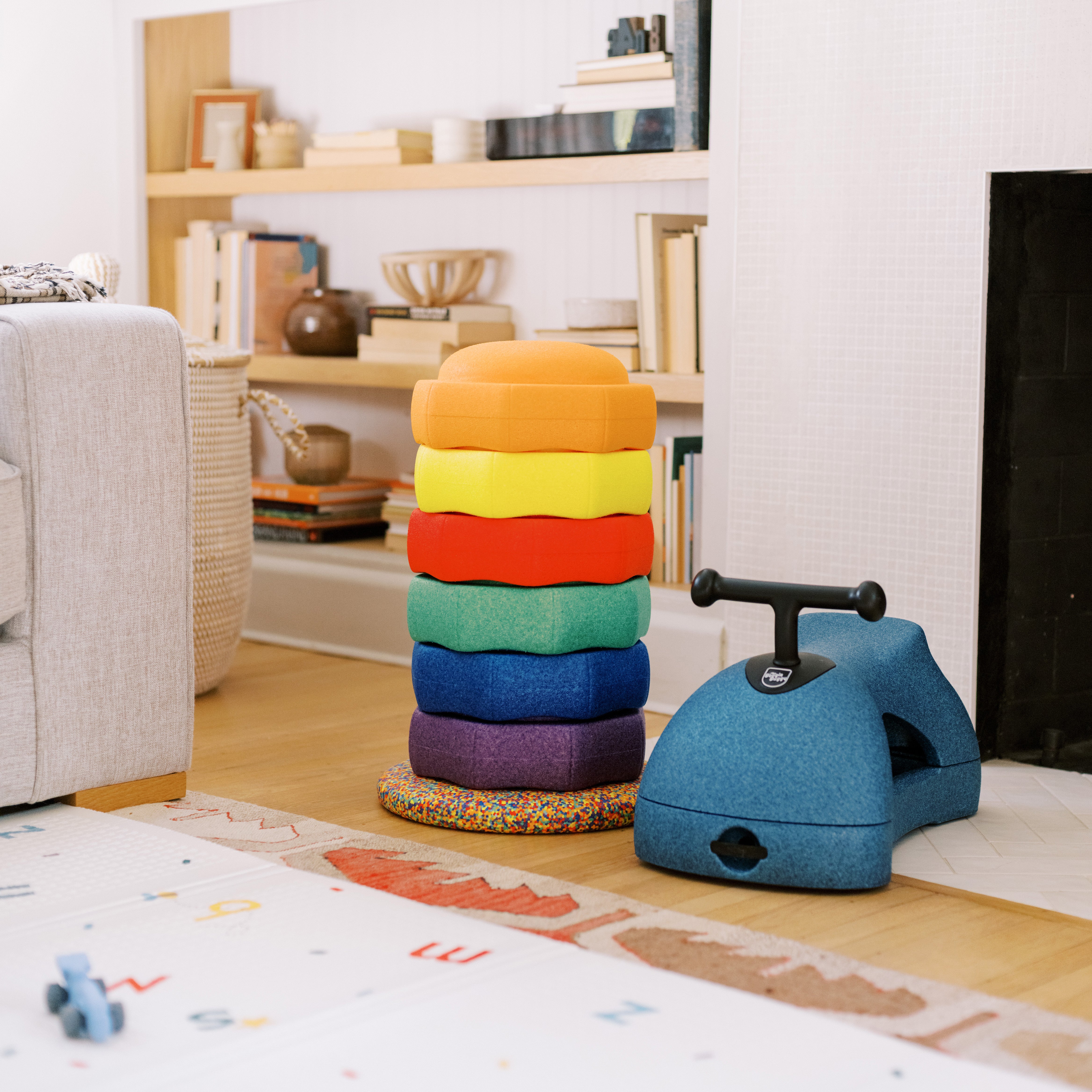 A stack of colorful, rounded EPP foam cushions arranged in a rainbow sits beside the Boogie Buggy | Blue scooter in a cozy living room with shelves, books, a gray couch, and a lettered play mat on the floor.