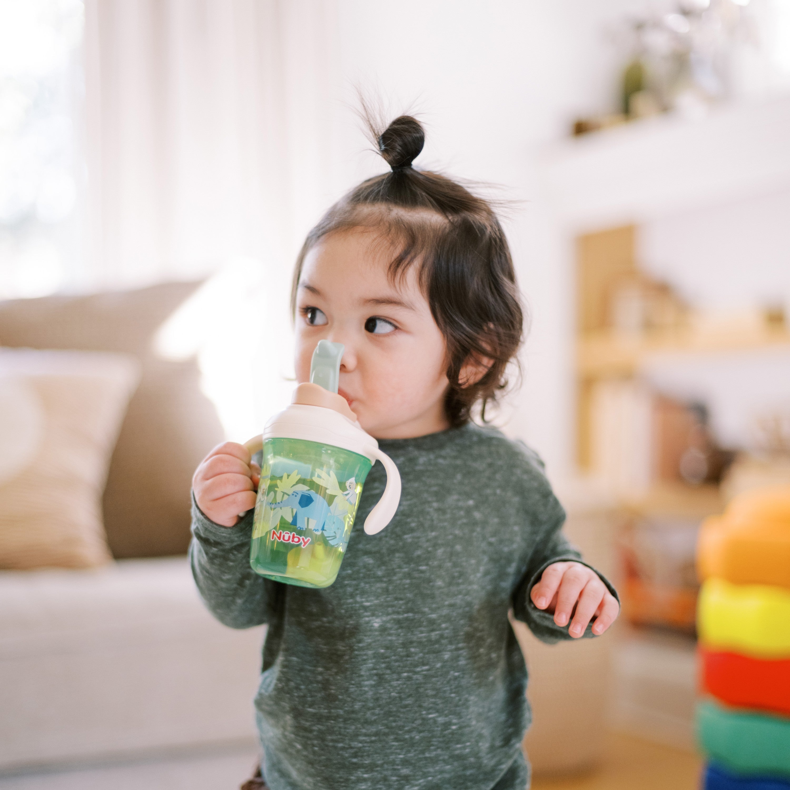 A dark-haired toddler with a small topknot drinks from a Nuby 2-in-1 Toddler Cup (Jungle & Ocean, 2 Pack) while standing in a bright, cozy living room with toys and shelves in the background.