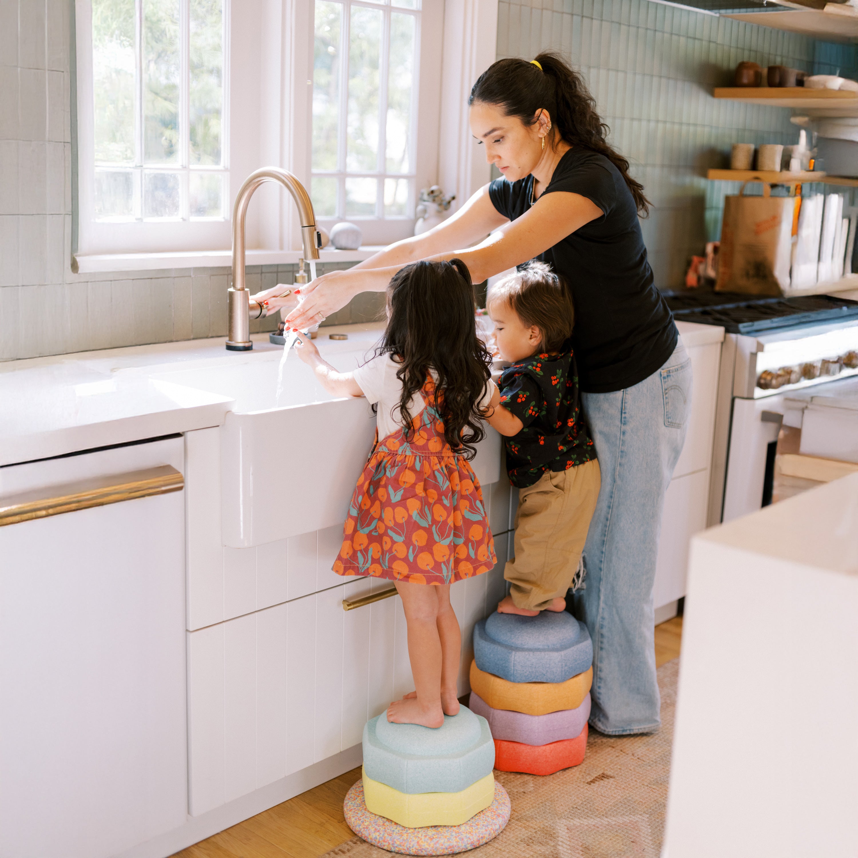 A woman helps two kids wash their hands at the kitchen sink. The children stand on StepPebbles and FREE Balance Board | Pastel, adding fun and balance to the bright, modern kitchen.