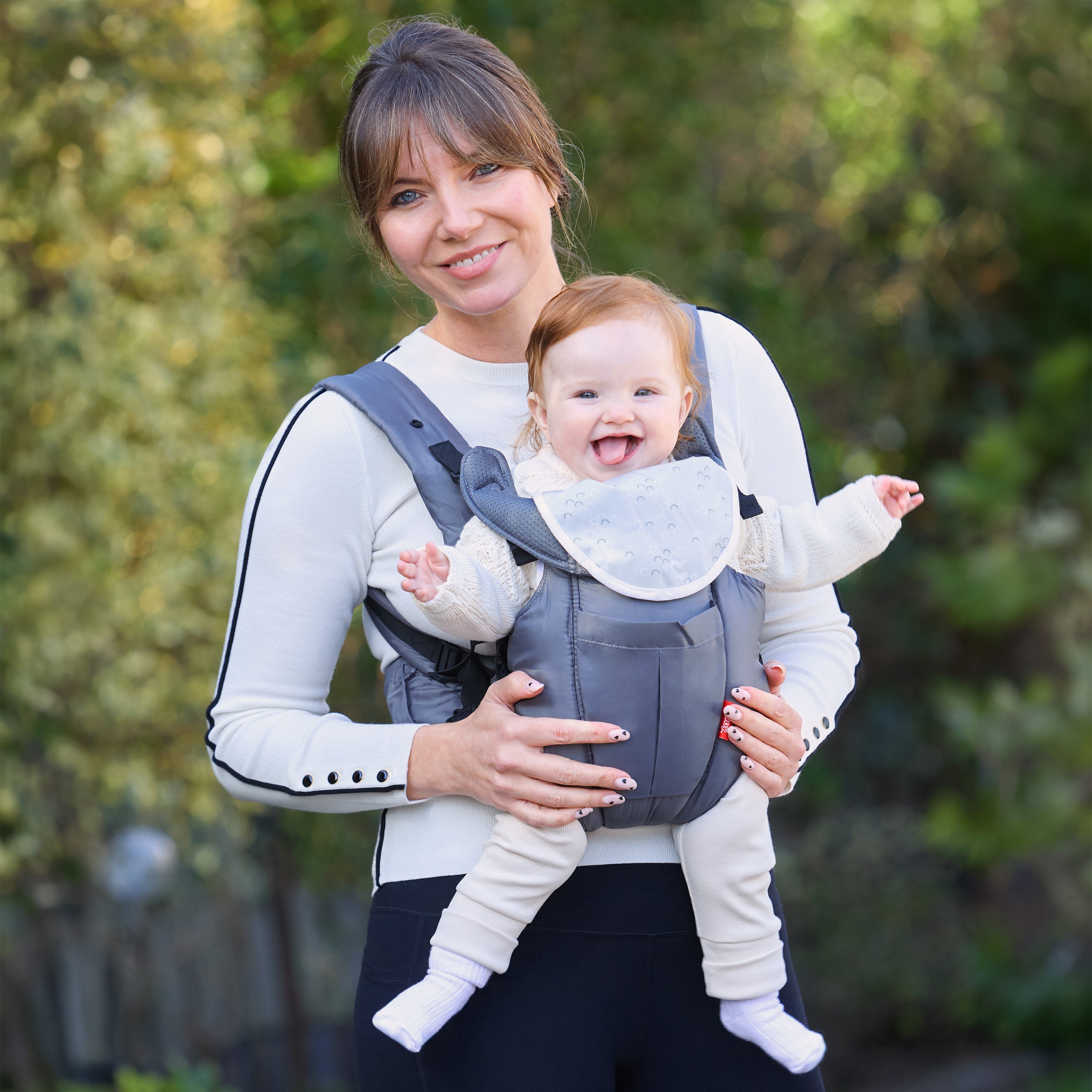 A smiling woman outdoors holds a happy baby in the Deluxe Baby Carrier | Grey. She wears a white sweater, and the lightly dressed baby with a bib is securely supported. A blurred background of greenery completes the scene.