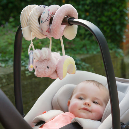 A baby sits in a car seat, gazing up at the Animal Adventures Spiral Activity Toy | Bunny in pink and cream, featuring a plush rabbit and pacifier attached, as it hangs from the car seat handle. Soft greenery is blurred in the background.