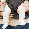 A person in blue jeans and a beige jacket holds a baby, dressed in white knitted pants and socks, secured in the SureFit Baby Carrier. Only the baby's legs and part of one hand are visible.