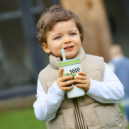 A young child with brown hair and blue eyes, dressed in a beige vest and white shirt, stands outside holding a Thirsty Kids Flexstraw Stainless Steel Cup | Monster Trucks. The background is blurred greenery and a building.