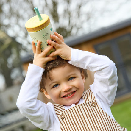 A young child with light brown hair and blue eyes lifts the Thirsty Kids Flexstraw Stainless Steel Cup | Dogs overhead, smiling. Dressed in a white shirt and tan striped overalls, they stand outside with blurred trees and a building behind them.
