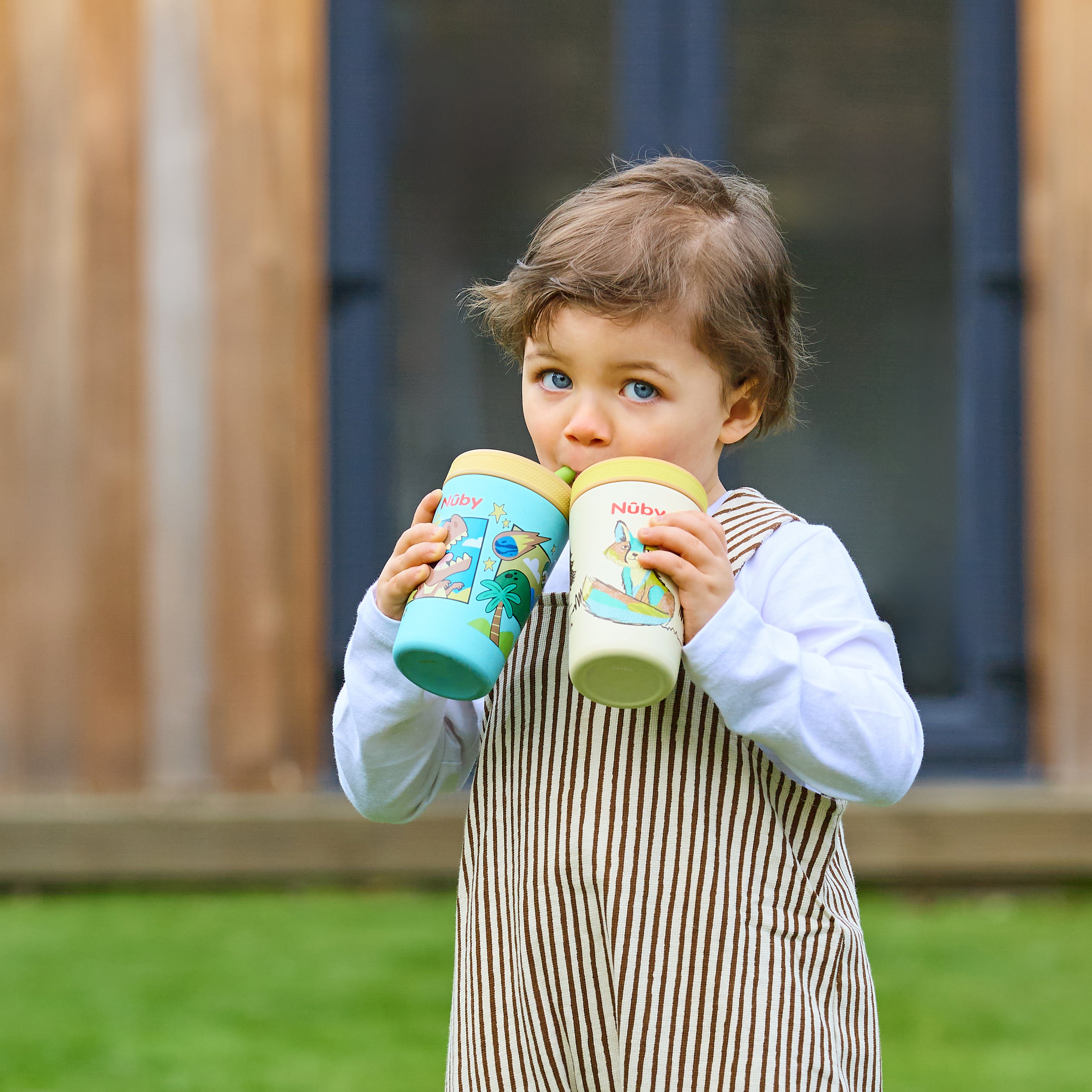 A young child with light brown hair and blue eyes stands outside, wearing a striped outfit while sipping from two Thirsty Kids Flexstraw Stainless Steel Cups | Dogs. The background shows blurred buildings and green grass.