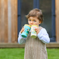 A young child with light brown hair and blue eyes stands outside, wearing a striped outfit while sipping from two Thirsty Kids Flexstraw Stainless Steel Cups | Dogs. The background shows blurred buildings and green grass.