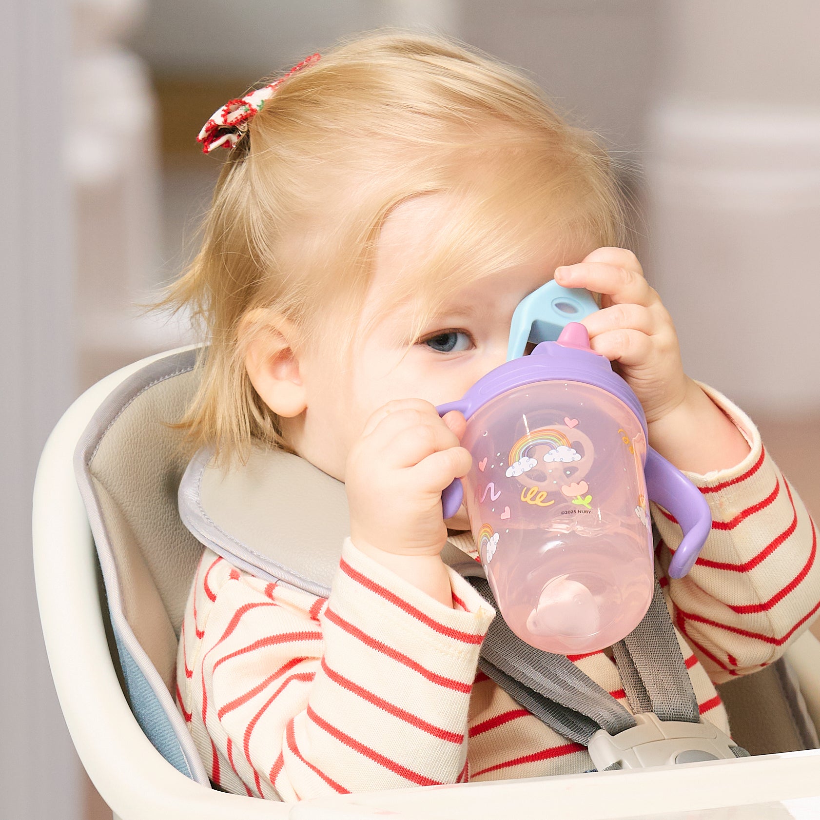 A young blonde child in a striped shirt with a red hair clip sits in a high chair, drinking from the 2-in-1 Toddler Cup (2 Pack) | Tiger & Rainbows.