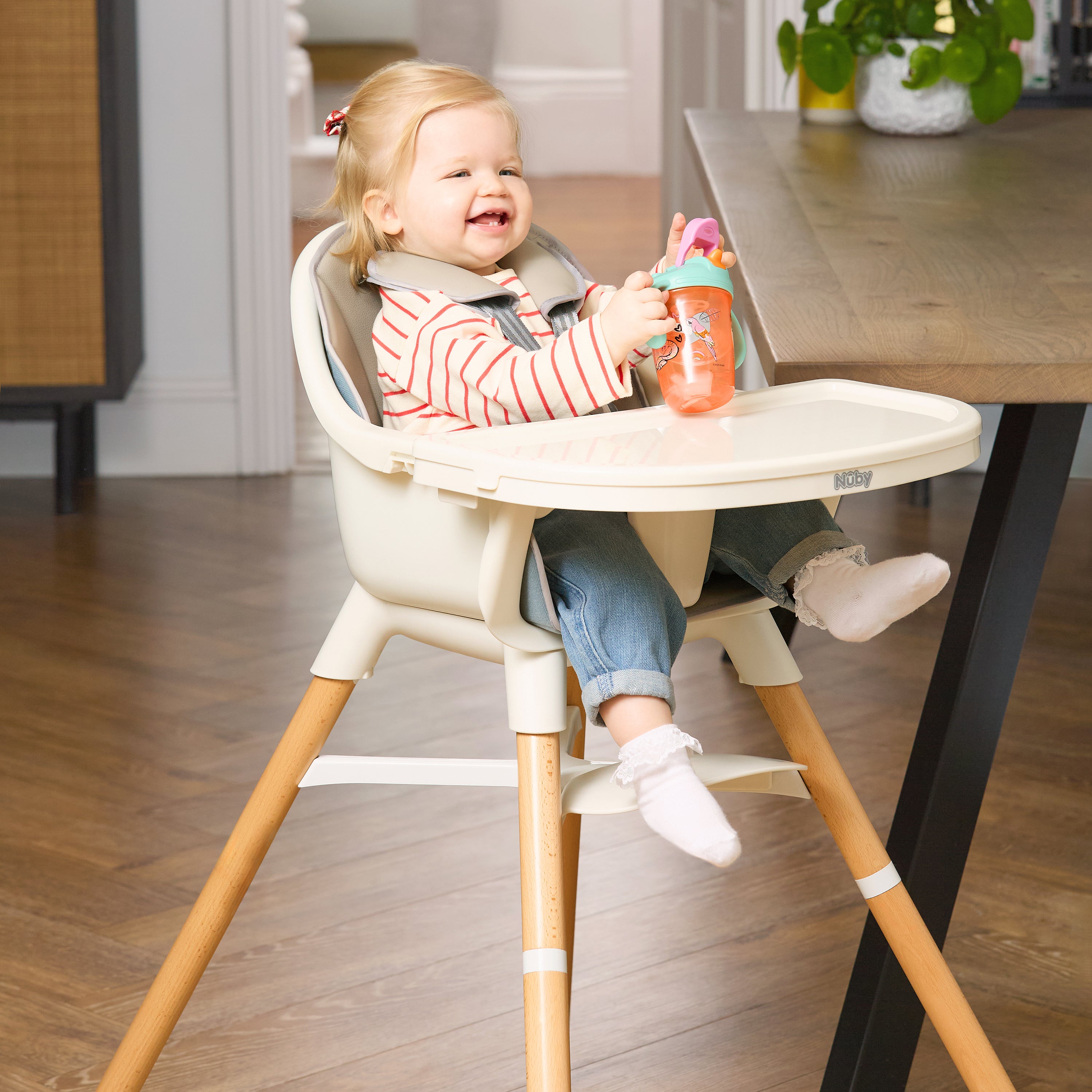 A smiling toddler with light hair sits at a wooden table in the 4-in-1 Multi-Use High Chair | Dark Beige, holding an orange sippy cup. The child wears a striped shirt and jeans, with wood floors and plants in the background.