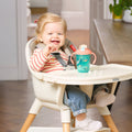 A smiling blonde toddler sits in a high chair, wearing a red and white striped shirt and blue jeans. On her tray is a Jungle & Ocean 2-in-1 Toddler Cup (2 Pack) with a pink lid and weighted straw.