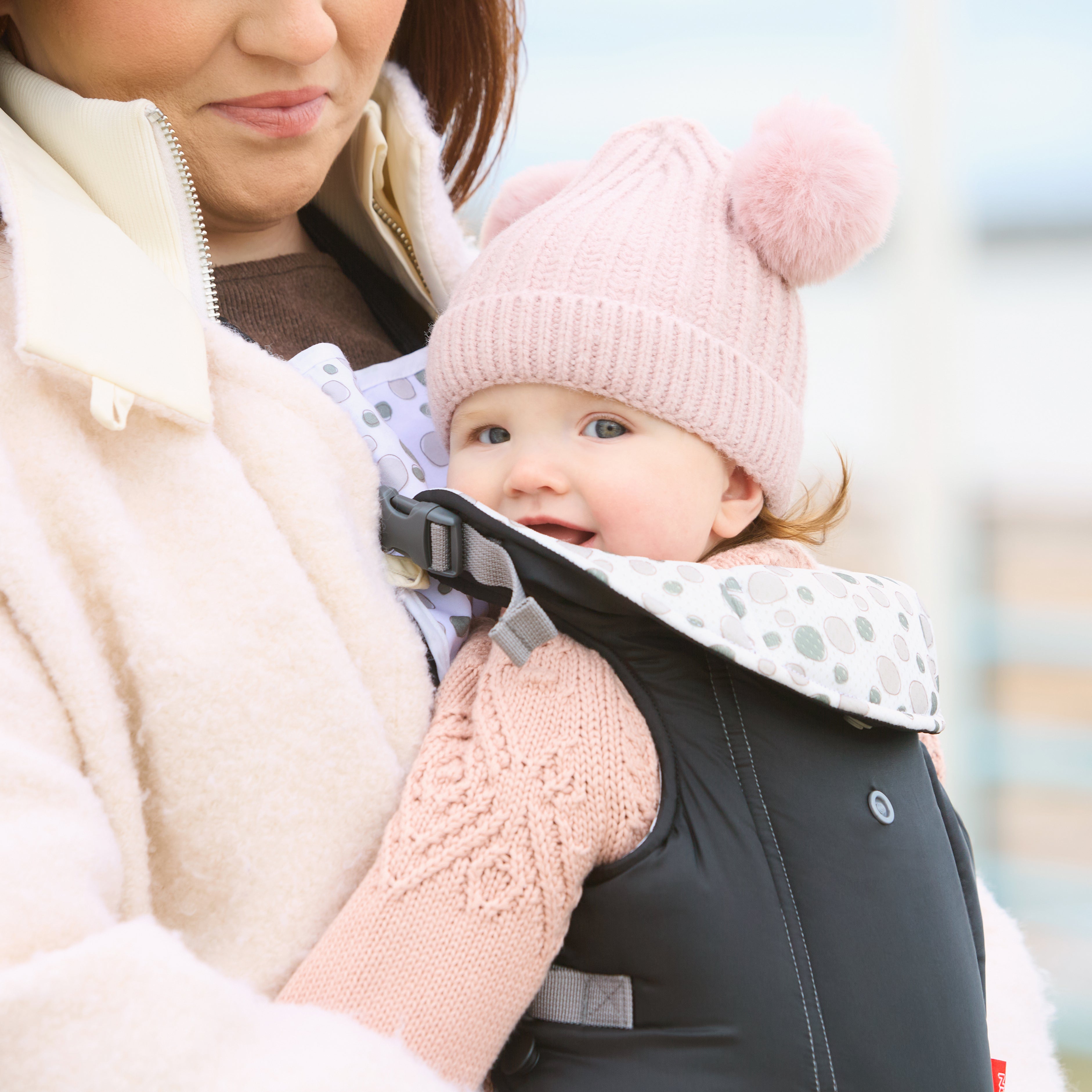 A woman carries a smiling baby in the Classic Baby Carrier | Black. The baby, dressed warmly in a pink knit pom-pom hat and matching sweater, enjoys the cool weather from a front-facing, breathable seat.