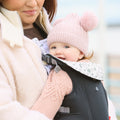 A woman carries a smiling baby in the Classic Baby Carrier | Black. The baby, dressed warmly in a pink knit pom-pom hat and matching sweater, enjoys the cool weather from a front-facing, breathable seat.