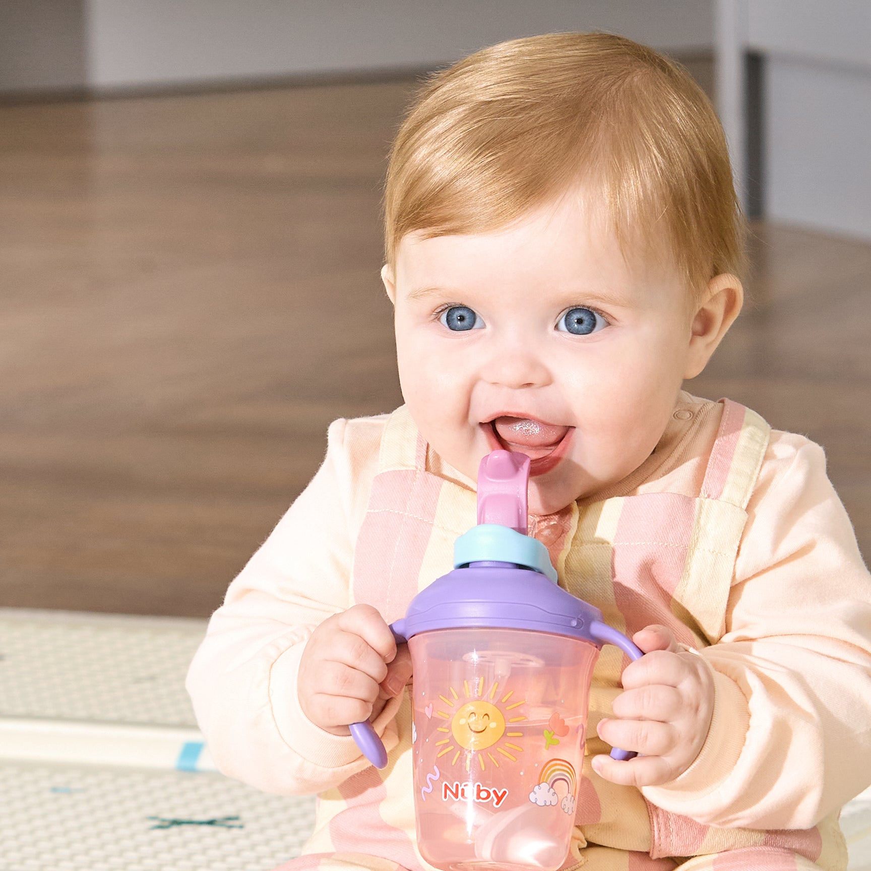 A baby with blonde hair and blue eyes smiles while drinking from a 2-in-1 Toddler Cup (2 Pack) | Tiger & Rainbows, featuring a weighted straw and colorful tiger and rainbow designs.