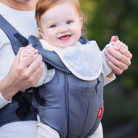 A smiling red-haired baby sits in the Deluxe Baby Carrier | Grey, wearing a patterned white bib. An adult in white holds the baby's hands, with softly blurred greenery in the background.