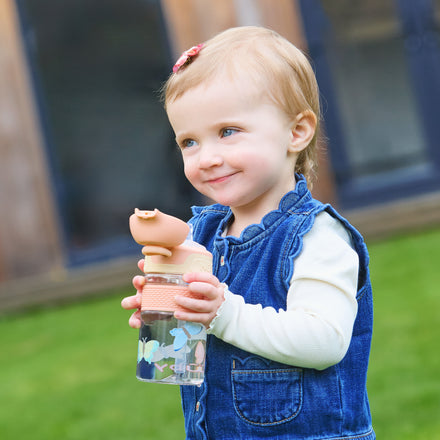 A smiling toddler with short blond hair, in a denim dress and white shirt, stands outdoors holding the Thirsty Kids Flip-It BOLT Water Bottle | Butterflies with a no-spill lid and colorful butterfly designs.