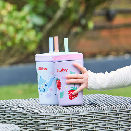A child’s hand holds a purple Thirsty Kids Flexstraw Stainless Steel Cup with strawberries and a green straw beside a blue whale-print cup, both on a wicker table outdoors.