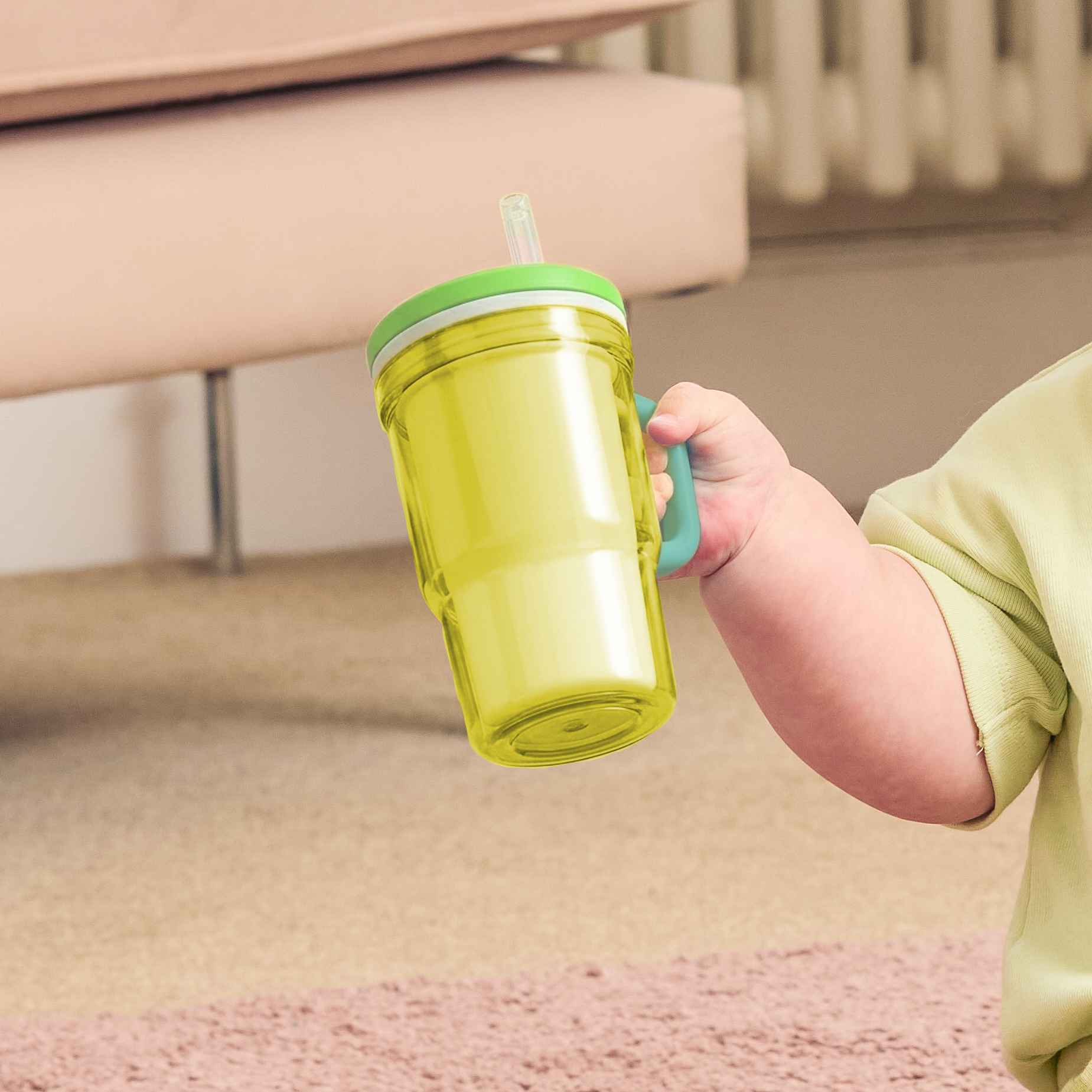 A child’s chubby arm holds the Insulated Tiny Tumbler | Lemon Lime—translucent yellow with a green lid, a small handle, and a silicone straw—set against a softly blurred indoor scene with beige carpet and a light pink sofa.