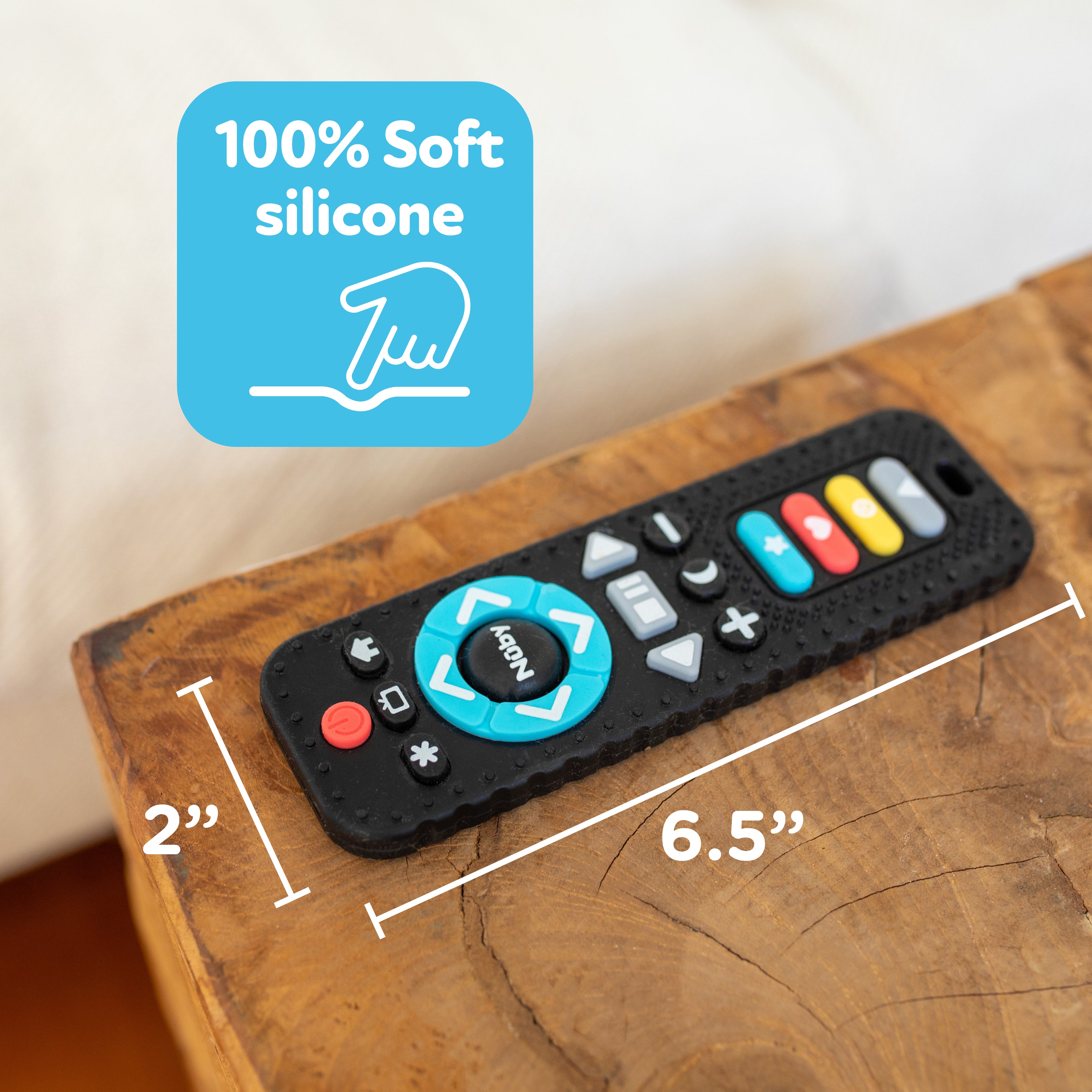 The Silicone Remote Control Teether Toy, made from 100% soft black silicone and measuring 6.5 inches by 2 inches, rests on a wooden table. A blue label featuring a hand icon highlights its gentle material, making it the perfect teething toy for babies.