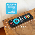 The Silicone Remote Control Teether Toy, made from 100% soft black silicone and measuring 6.5 inches by 2 inches, rests on a wooden table. A blue label featuring a hand icon highlights its gentle material, making it the perfect teething toy for babies.