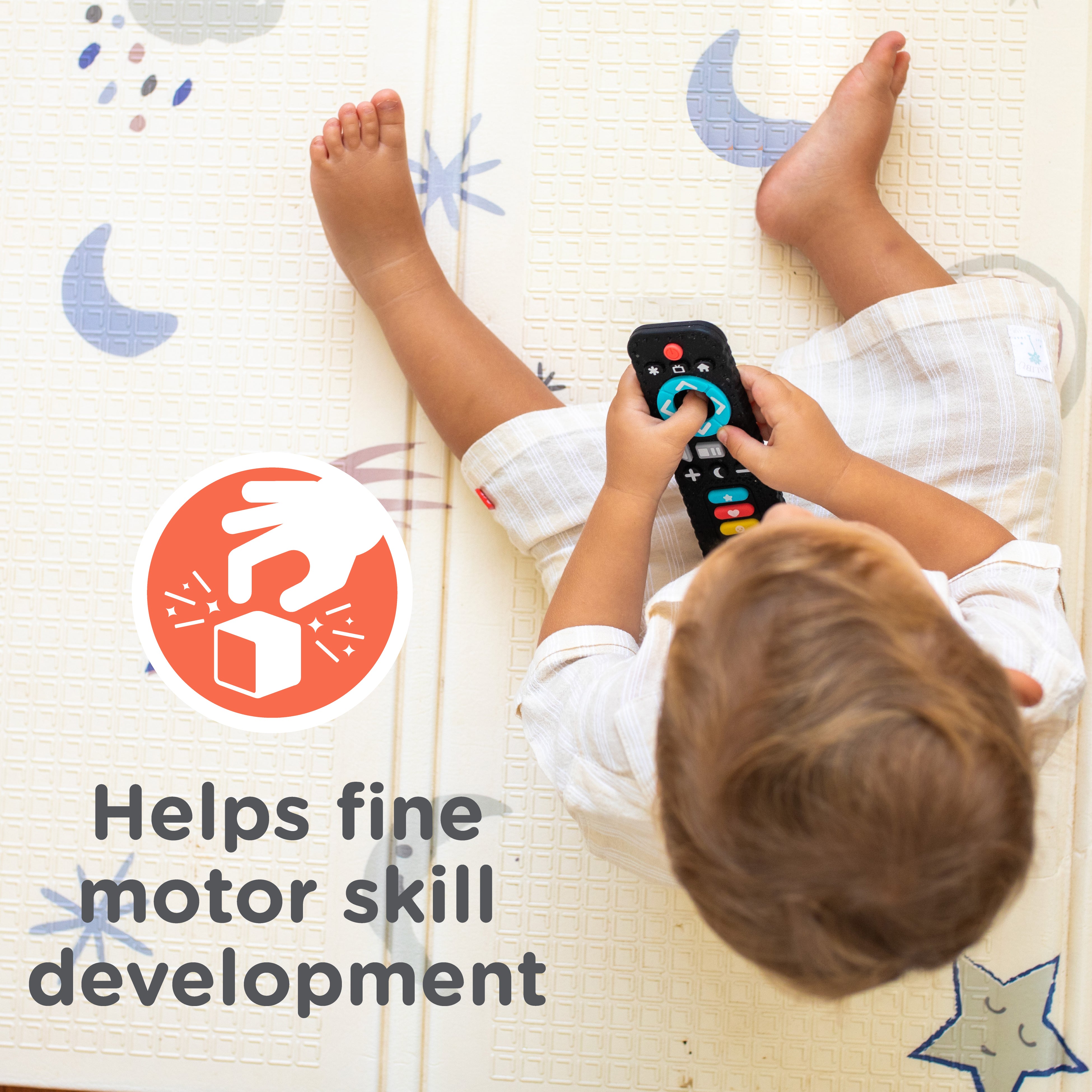 A toddler wearing white clothing sits on a play mat, holding the Silicone Remote Control Teether Toy. Text reads, “Helps fine motor skill development” next to an icon of a hand manipulating a block.
