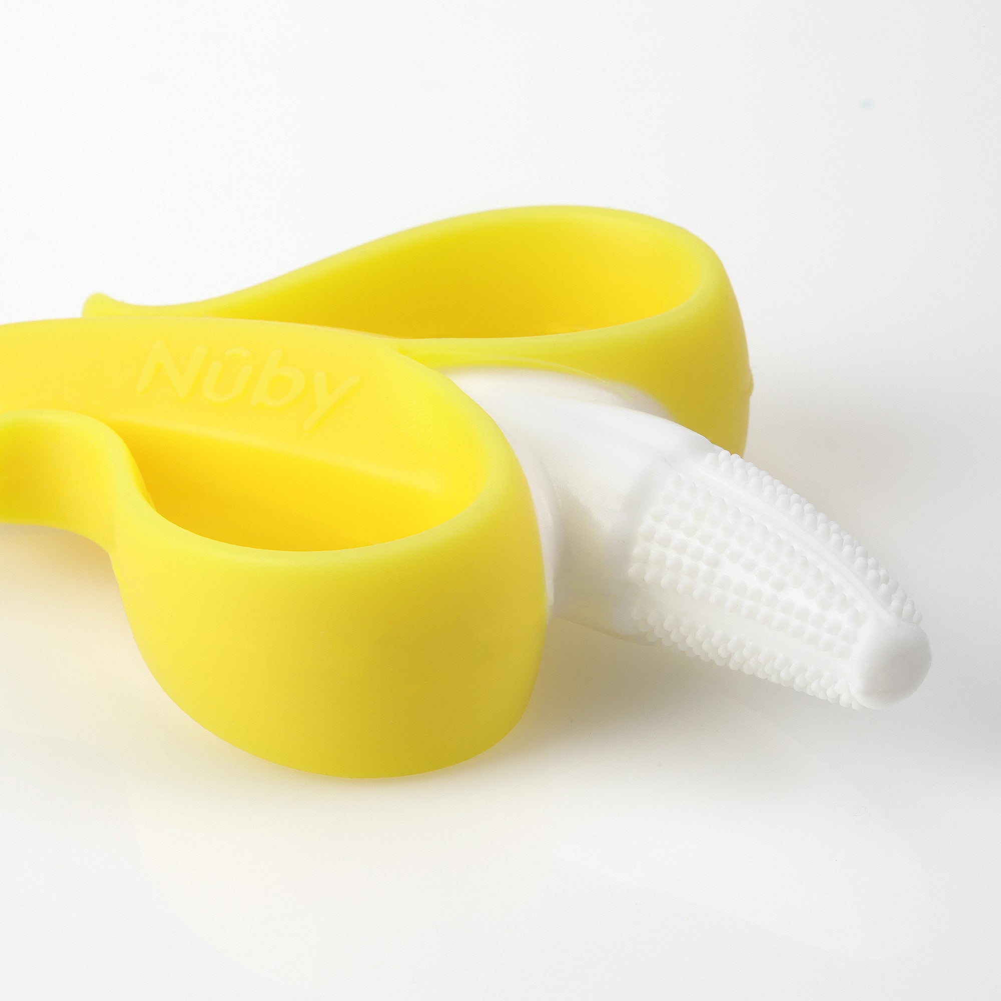 A NanaNubs Banana Massaging Toothbrush, made of soft yellow and white silicone and shaped like a peeled banana, lies on a white surface.