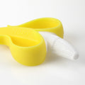 A NanaNubs Banana Massaging Toothbrush, made of soft yellow and white silicone and shaped like a peeled banana, lies on a white surface.