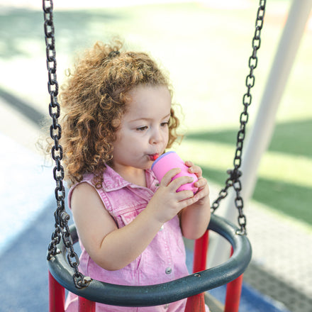 A young child with curly hair sits on a playground swing, wearing a pink sleeveless shirt and drinking from the Nuby US 3-Stage Training Cup Set in Aqua. Blurred playground equipment and green grass are visible in the background.