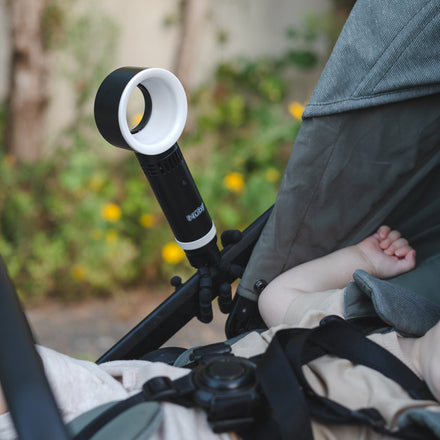 A Nuby Bladeless Fan is clipped to a stroller, gently blowing air toward a resting baby whose arm and hand are visible, with greenery and softly blurred yellow flowers in the background.