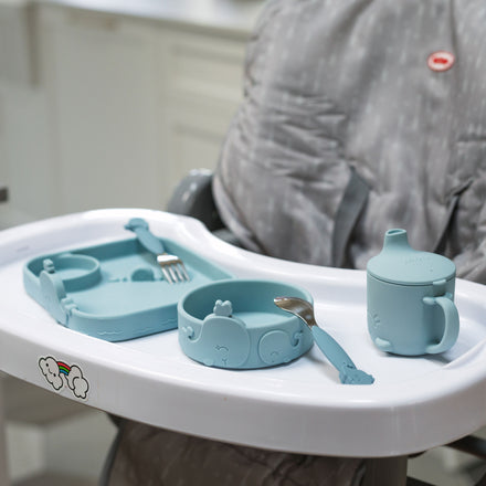 A Nuby Animal Friends 6-Piece Dinnerware Set - Blue Whale sits on a baby high chair with a white tray and gray patterned cover. The blue silicone set includes a plate, bowl, fork, spoon, sippy cup, and features a playful whale design.