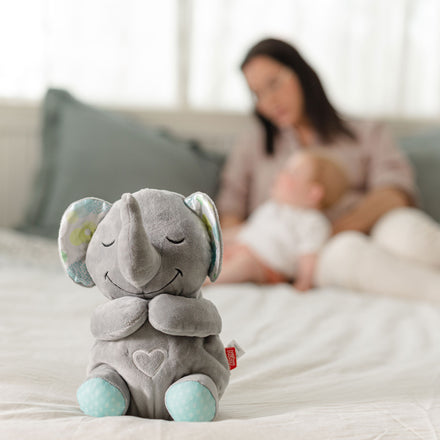 A Motion & Sound Plush in soft gray, shaped like an elephant, sits on a bed in focus while a woman holds a baby in the blurred background, both resting against pillows.