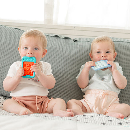 Two baby twins sit on a gray couch, each holding a Nuby US Pretend Play Teething Bundle silicone teether for soothing relief. They wear matching light shirts and peach shorts, with curious expressions. Natural light from the window behind them.