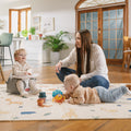 A woman on a Nuby playmat with two young children. One child is crawling toward Nuby toys. The other is sitting in a Nuby My Floor Seat with Activity Tray.